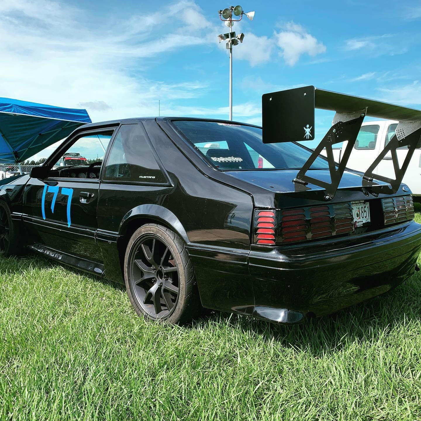 A black sports car with the number 177 in blue on its side is parked on a grassy field. The car, featuring the Mustang Big Wang Kit '79-‘93 Fox body by 9livesracing for optimal down-force, boasts aftermarket rims and a large rear spoiler. A partly cloudy sky and other vehicles are visible in the background, highlighting its impressive lift-to-drag ratio.