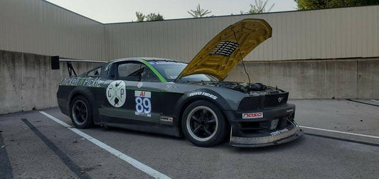 A black race car with the number 89 on its door sits parked with its hood open. The car boasts various sponsor logos and prominently features a Mustang Big Wang Kit '05-09 S197 from 9livesracing as its performance rear wing. It is parked in an outdoor lot with barriers and a metal building in the background.