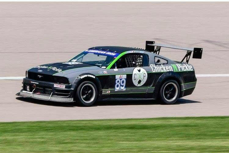 A black race car with the number 89 on the door, adorned with the "Chicken N Pickle" logo and graphics, speeds down a racetrack. The car is equipped with a 9livesracing Mustang Big Wang Kit '05-09 S197 aluminum wing for superior downforce adjustments and features various sponsor decals. Grass is visible in the foreground.