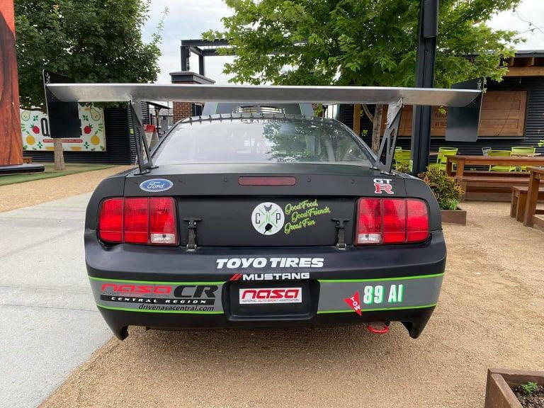 A black Ford Mustang race car adorned with decals such as "TOYO TIRES," "NASA," and "89 AI" is parked outdoors. Equipped with the 9livesracing Mustang Big Wang Kit '05-09 S197 for optimal downforce adjustments, the car also features a message on the trunk reading "Good Food Good Friends Good Times." The backdrop includes trees, a picnic area, and modern buildings.