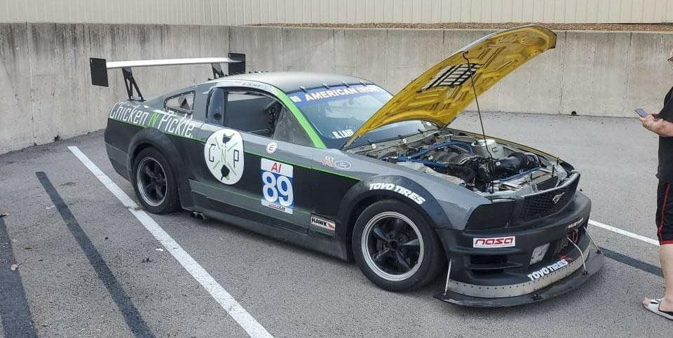 A race car with number 89 displayed on the side is parked in a lot, its hood open to reveal the engine. It boasts features like the Mustang Big Wang Kit '05-09 S197 by 9livesracing for optimal downforce adjustments, along with "American Iron" and various sponsor decals. Nearby, a person is holding a mobile phone.