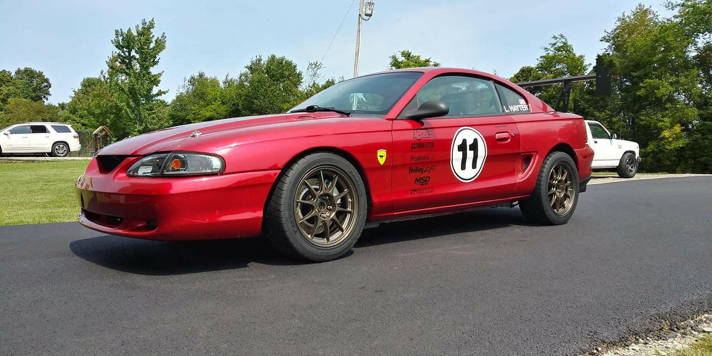 A red Mustang Big Wang Kit '94-‘98 SN95 from 9livesracing, adorned with a black number "11" in a white circle decal on the door and several other racing decals, bronze wheels, and extruded aluminum wings, is parked on a paved driveway. In the background, a white vehicle is visible next to a grassy area and trees.