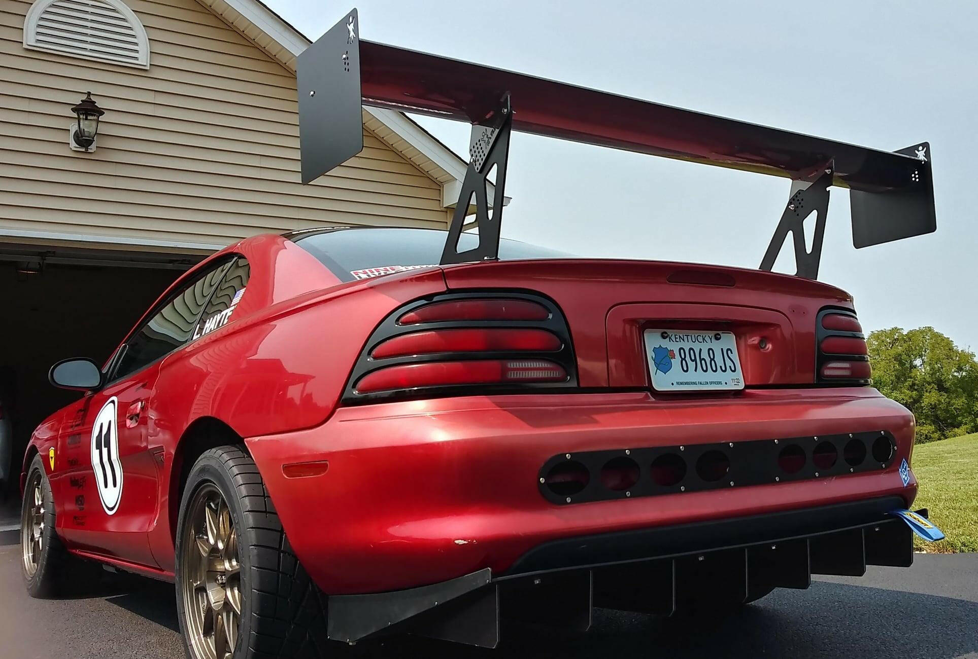 A Mustang equipped with the 9livesracing Big Wang Kit for '94-‘98 SN95, featuring a large rear wing, black rear diffuser, extruded aluminum wings, a number #8 decal on the side, and bronze-colored wheels is parked on a driveway in front of a garage. The rear license plate reads "9848JS," showcasing its aerodynamic design for an optimal lift-to-drag ratio.