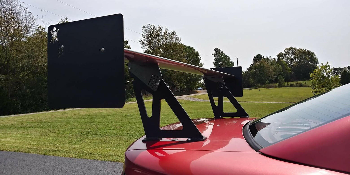 A close-up of a Mustang Big Wang Kit '94-'98 SN95 by 9livesracing, featuring extruded aluminum wings attached to the trunk of a red sports car. The background showcases a lawn, trees, and a paved driveway under a cloudy sky.