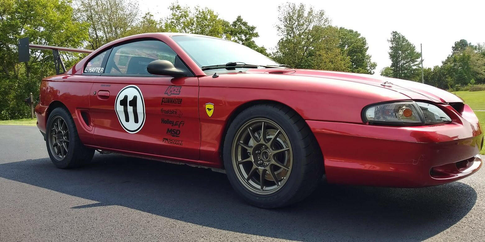 A red sports car with the number 11 displayed on the side is parked on an asphalt surface, showcasing various logos and decals. The vehicle features bronze-colored alloy wheels and is fitted with a 9livesracing Mustang Big Wang Kit '94-'98 SN95, characterized by its extruded aluminum wing. Trees and grass can be seen in the background under a clear sky.