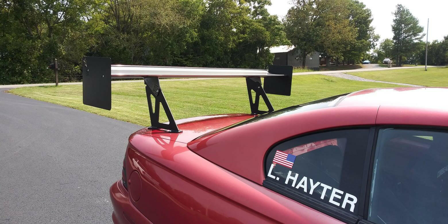 A red sports car parked beside a grassy area with trees in the background. The car boasts a Mustang Big Wang Kit '94-‘98 SN95 rear wing spoiler by 9livesracing and an American flag decal on the rear side window, displaying the name "L. HAYTER." The vehicle sits on a smooth, paved surface, optimized for lift-to-drag ratio efficiency.