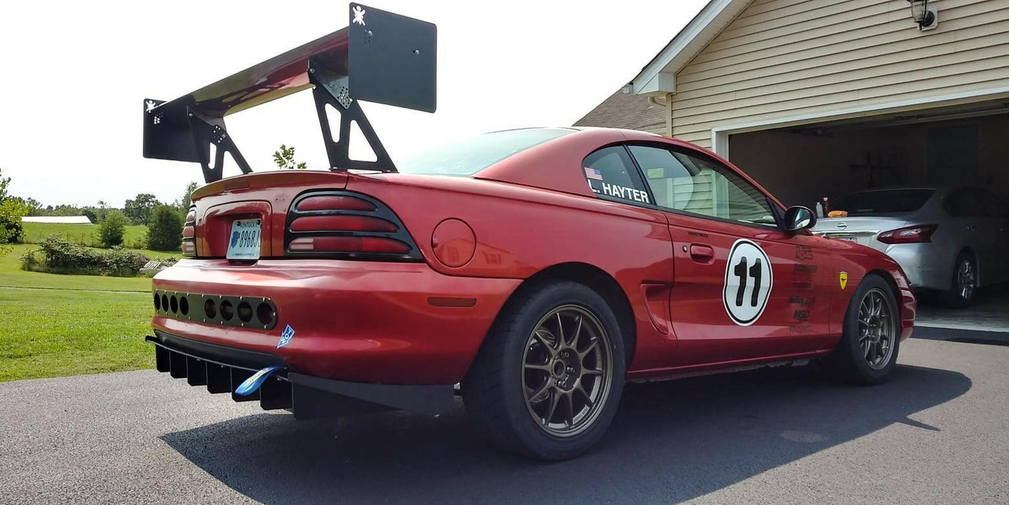 A red race car with a large rear spoiler and the number 77 on the door is parked in front of a garage. The car features various decals and modifications, including racing wheels, USA flag, blue tow hooks, and an impressive Mustang Big Wang Kit '94-‘98 SN95 by 9livesracing for optimal lift to drag ratio. Another vehicle and a beige house are visible in the background.