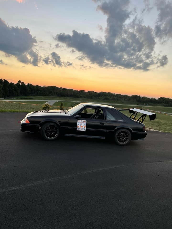 A Mustang Big Wang Kit '79-‘93 Fox body by 9livesracing, featuring a sleek black finish and a prominent rear wing for optimal down-force along with the racing number “717” on the door, is parked on a paved surface. The background showcases a grassy field and a picturesque sunset with scattered clouds in the sky.