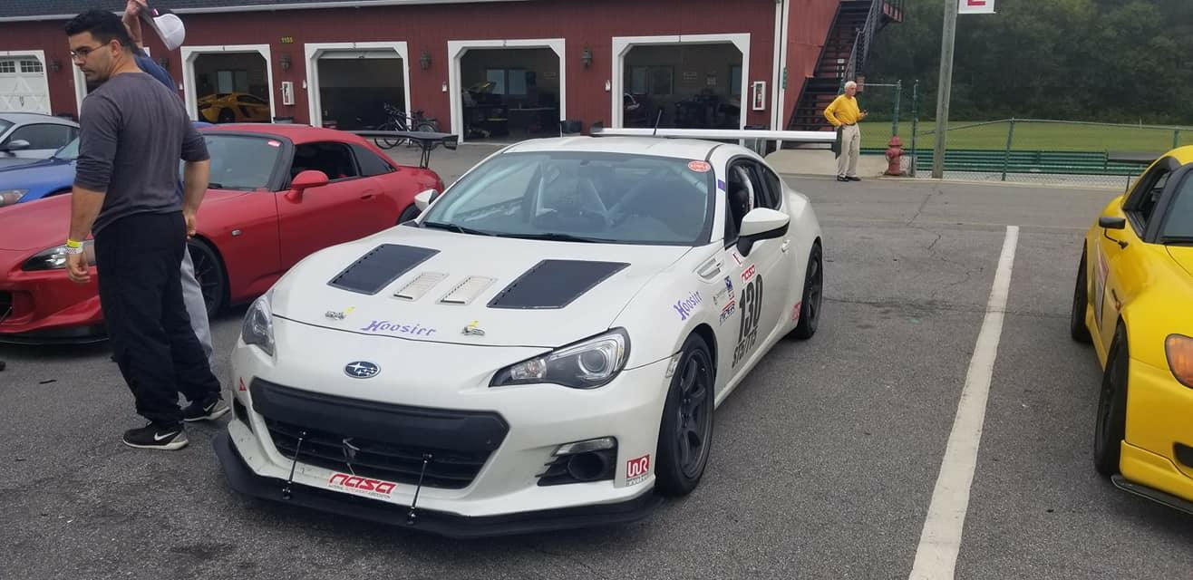 A white Subaru sports car, enhanced with a GT86 Big Wang Kit '12-'21 FRS / BRZ / 86 from 9livesracing, adorned with various decals and modifications including extruded aluminum wings for an improved lift-to-drag ratio, is parked in a lot among other cars, with a red one to the left and a yellow one to the right. Several people are standing around, and a red building with open garages is visible in the background.