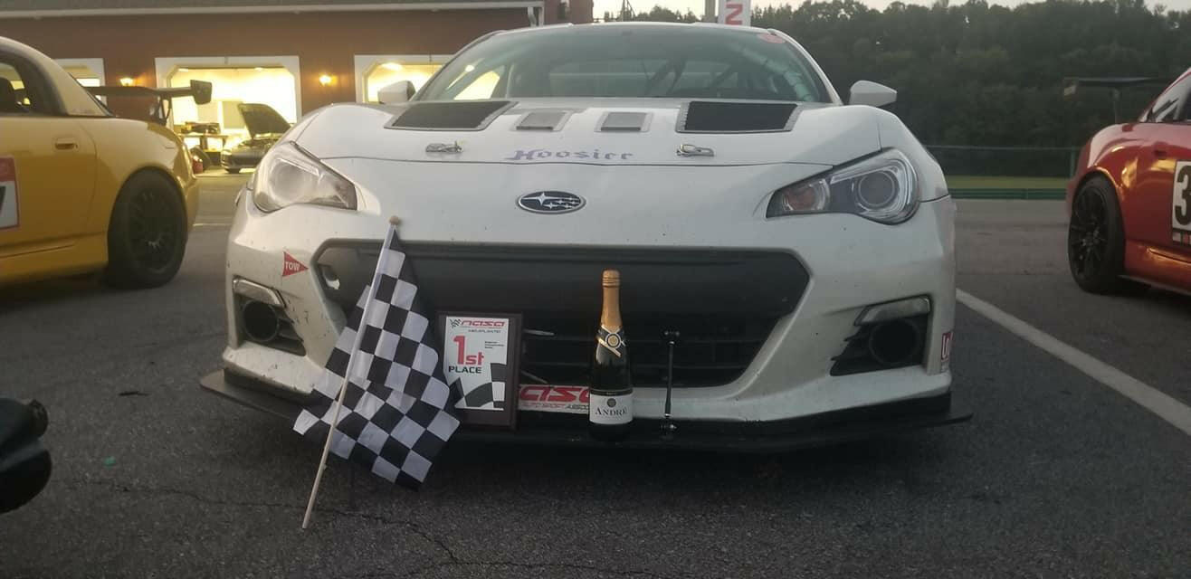 Front view of a white race car with a Subaru logo and the word "Hoosier" on the hood, proudly displaying a first-place plaque, a checkered flag, and a bottle of champagne on the front bumper. Equipped with the GT86 Big Wang Kit '12-'21 FRS / BRZ / 86 from 9livesracing for exceptional performance featuring extruded aluminum wings, it is parked alongside yellow and red cars in the lot.