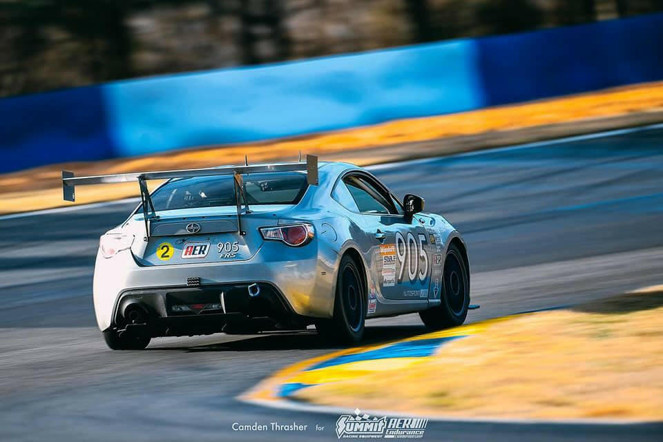 A sleek silver race car, labeled with the number 905, speeds on a race track. The car, featuring green and white livery and visible sponsor logos, boasts the 9livesracing GT86 Big Wang Kit for '12-'21 FRS / BRZ / 86 models with extruded aluminum wings for optimal lift-to-drag ratio. Trees and a blue barrier line the track in the background. Shot by Camden Thrasher for Yummi AER.