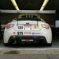 A white GT86 equipped with the 9livesracing Big Wang Kit, featuring the number 130 on its back, is parked in a garage. The car displays various stickers such as "NASA," "Hoosier," and "ST5," and boasts extruded aluminum wings for a superior lift-to-drag ratio. Two motorcycles are partially visible inside the garage.