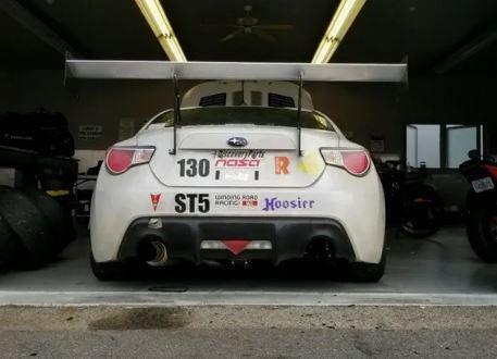 A white GT86 equipped with the 9livesracing Big Wang Kit, featuring the number 130 on its back, is parked in a garage. The car displays various stickers such as "NASA," "Hoosier," and "ST5," and boasts extruded aluminum wings for a superior lift-to-drag ratio. Two motorcycles are partially visible inside the garage.