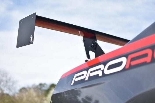 Close-up of the rear wing, specifically the Mustang Big Wang Kit '10-14 S197 II by 9livesracing, featuring an extruded aluminum performance airfoil, on a black and red sports car with the word "PROA" partially visible on the side, set against a blurred background of trees and a cloudy sky.