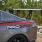 Close-up of a gray sports car's rear, featuring the 9livesracing Mustang Big Wang Kit '10-14 S197 II with adjustable downforce, black alloy wheels, and a "PROAUTO" decal on the side. The background shows green trees and a rocky area.