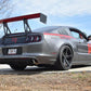 A sleek gray sports car with a large rear wing and black alloy wheels is parked on a road. Equipped with the Mustang Big Wang Kit '10-14 S197 II by 9livesracing, which includes extruded aluminum wings for downforce adjustment, it prominently features a racing number "14" on the side and the text "PROAUTO" on the rear window. Trees and a house can be seen in the background under a partly cloudy sky.