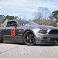 A gray Mustang Big Wang Kit '10-14 S197 II, proudly displaying the number 14 in red on its side, is parked on a road. This car from 9livesracing boasts various decals, black rims, and a prominent front splitter with an extruded aluminum wing designed for optimal downforce adjustment. In the background, trees and a patch of blue sky with some clouds can be seen.