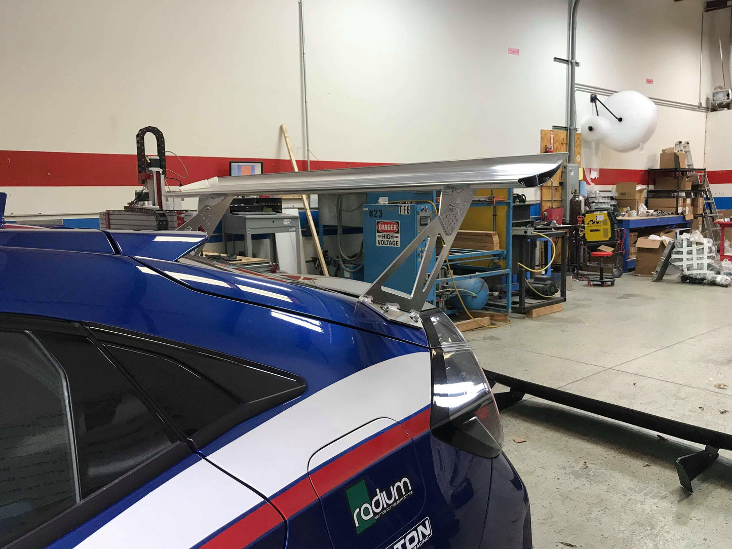 A blue sports car featuring a striking 9livesracing Civic Type-R Wang kit 2017+ FK8 rear wing spoiler is parked in a garage. The interior of the garage is filled with various tools, equipment, and a workspace with shelves and boxes in the background. The car has decals and logos on the side, showcasing modifications and sponsors aimed at optimizing down-force.