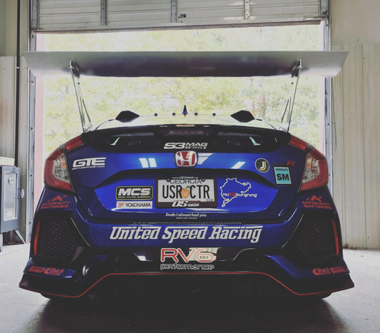 A blue racing car adorned with numerous sponsors' stickers, including "United Speed Racing" and a prominent "Honda" badge, features a large Big Wang GT3 rear wing from the Civic Type-R Wang kit 2017+ FK8 by 9livesracing. The car is parked in a garage with partially open doors, revealing trees outside.