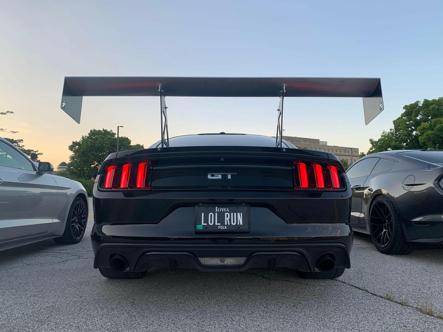 A black Mustang, fitted with the 9livesracing Big Wang Kit '14+ S550 aluminum wings to boost its down-force, is parked in a lot. The license plate reads "LOL RUN." Two other cars are partially visible on either side of the black Mustang. Trees and a building are in the background under an evening sky.