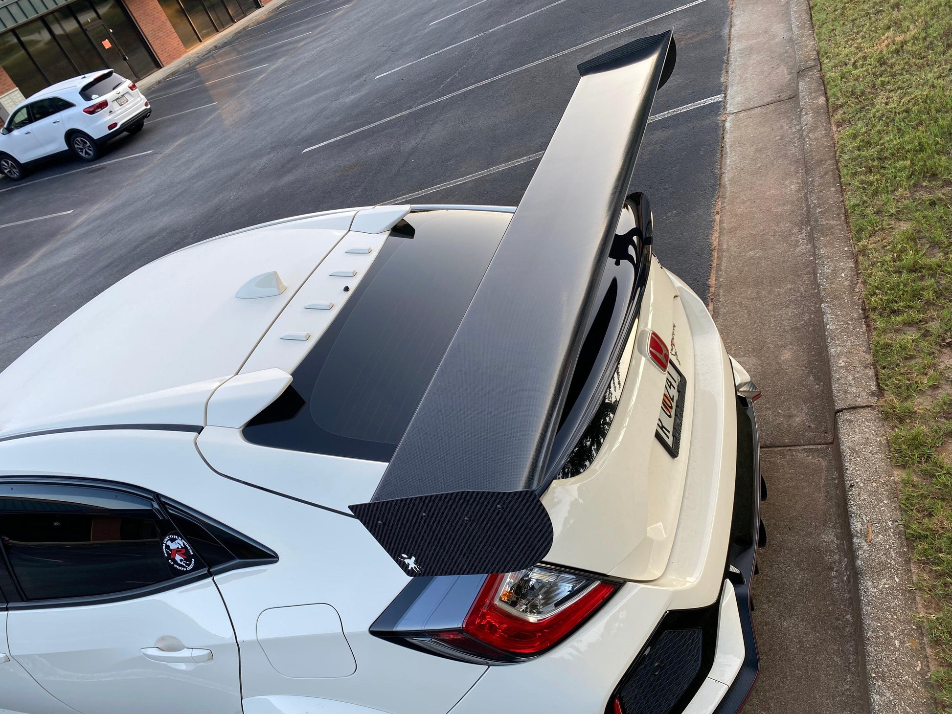 A white car with a large 9livesracing Civic Type-R Wang kit 2017+ FK8 carbon fiber rear spoiler is parked near a curb in a mostly empty parking lot. The car features black accents, including a prominent rear diffuser and window visors, optimizing its lift-to-drag ratio for increased down-force. The surrounding area shows some grass and a nearby building.