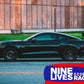 A sleek, black sports car with aluminum wings is parked on the street in front of a building with large windows. The prominent rear spoiler enhances its Mustang performance, showcasing the Mustang Big Wang Kit '14+ S550 by 9livesracing. The 9livesracing logo, featuring blue and red colors with a small cat graphic, is displayed in the bottom right corner.