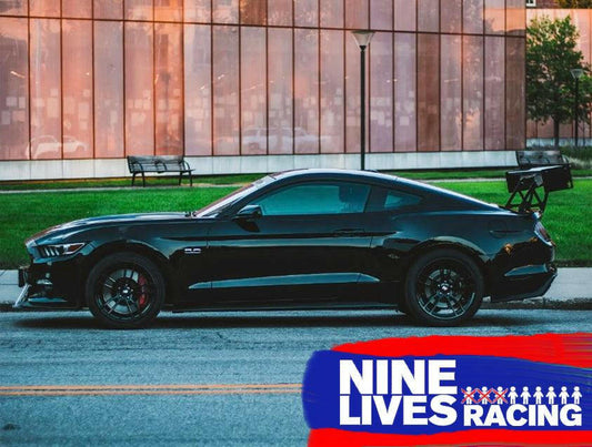 A sleek, black sports car with aluminum wings is parked on the street in front of a building with large windows. The prominent rear spoiler enhances its Mustang performance, showcasing the Mustang Big Wang Kit '14+ S550 by 9livesracing. The 9livesracing logo, featuring blue and red colors with a small cat graphic, is displayed in the bottom right corner.