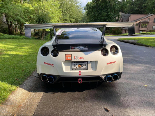 A white sports car with a large rear spoiler, specifically featuring the 9livesracing GT-R Big Wang Kit '07-22 for optimal down-force adjustments, parked on a driveway. The car has several decals on the back, including "Yokohama," "Tire Rack," and "G-LOC." The license plate reads "RC72896." Houses and trees dot the background.