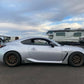 A sleek silver GR86 / BRZ fitted with bronze wheels is parked on an asphalt surface. The 9livesracing Extruded Aluminum Wings enhance its sporty appeal. In the background, there are a few other cars and a large camper under a partly cloudy sky.