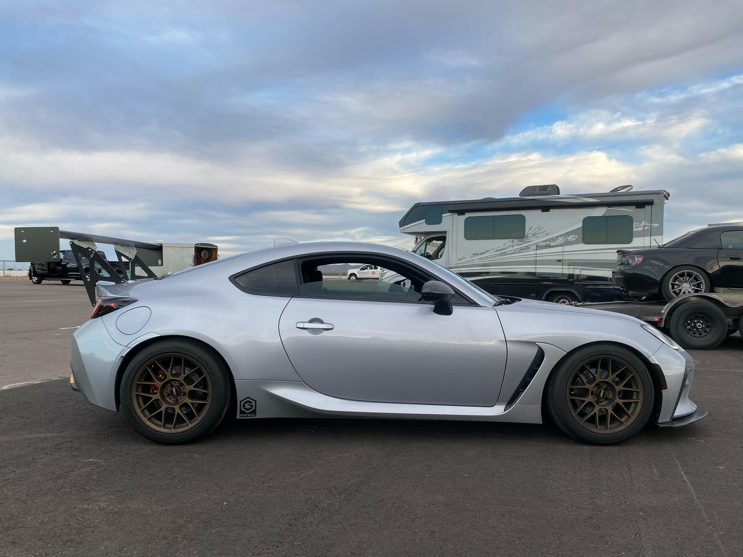 A silver sports car, parked in an open lot under a partly cloudy sky, boasts bronze rims and a sleek design. It features the GR86 / BRZ Wang Kit '22+ by 9livesracing with extruded aluminum wings, hinting that it may be modified for racing. In the background, there is a large RV and other parked vehicles, including another race car on a trailer.