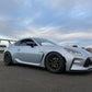 A sleek silver GR86 equipped with the 9livesracing Wang Kit '22+ featuring bronze alloy wheels is parked on an asphalt surface under a partly cloudy sky. The car boasts extruded aluminum wings that enhance its performance airfoils and a contoured body design. Other cars and objects are blurred in the background, emphasizing its standout features.