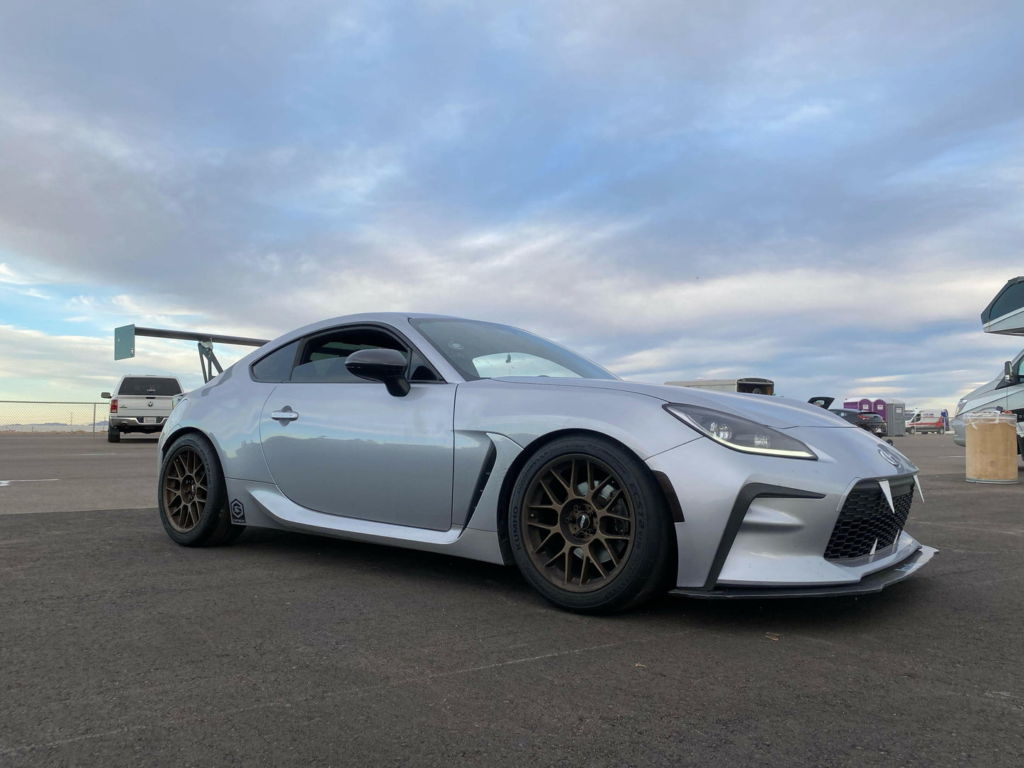 A sleek silver GR86 equipped with the 9livesracing Wang Kit '22+ featuring bronze alloy wheels is parked on an asphalt surface under a partly cloudy sky. The car boasts extruded aluminum wings that enhance its performance airfoils and a contoured body design. Other cars and objects are blurred in the background, emphasizing its standout features.