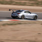 A silver sports car equipped with the 9livesracing GR86/BRZ Wang Kit '22+ and extruded aluminum wings is speeding on a racetrack. The car leans slightly as it navigates a turn, with blurred surroundings indicating high speed. The tarmac is bordered by red, white, and blue stripes, contrasting against the sandy landscape in the background.