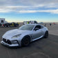 A silver sports car equipped with a 9livesracing GR86 / BRZ Wang Kit '22+, boasting extruded aluminum wings, is parked on a racetrack next to a black race car sporting the number 14. Several people and vehicles are in the background under a cloudy sky, creating an atmosphere of what seems to be a motorsport event or track day.