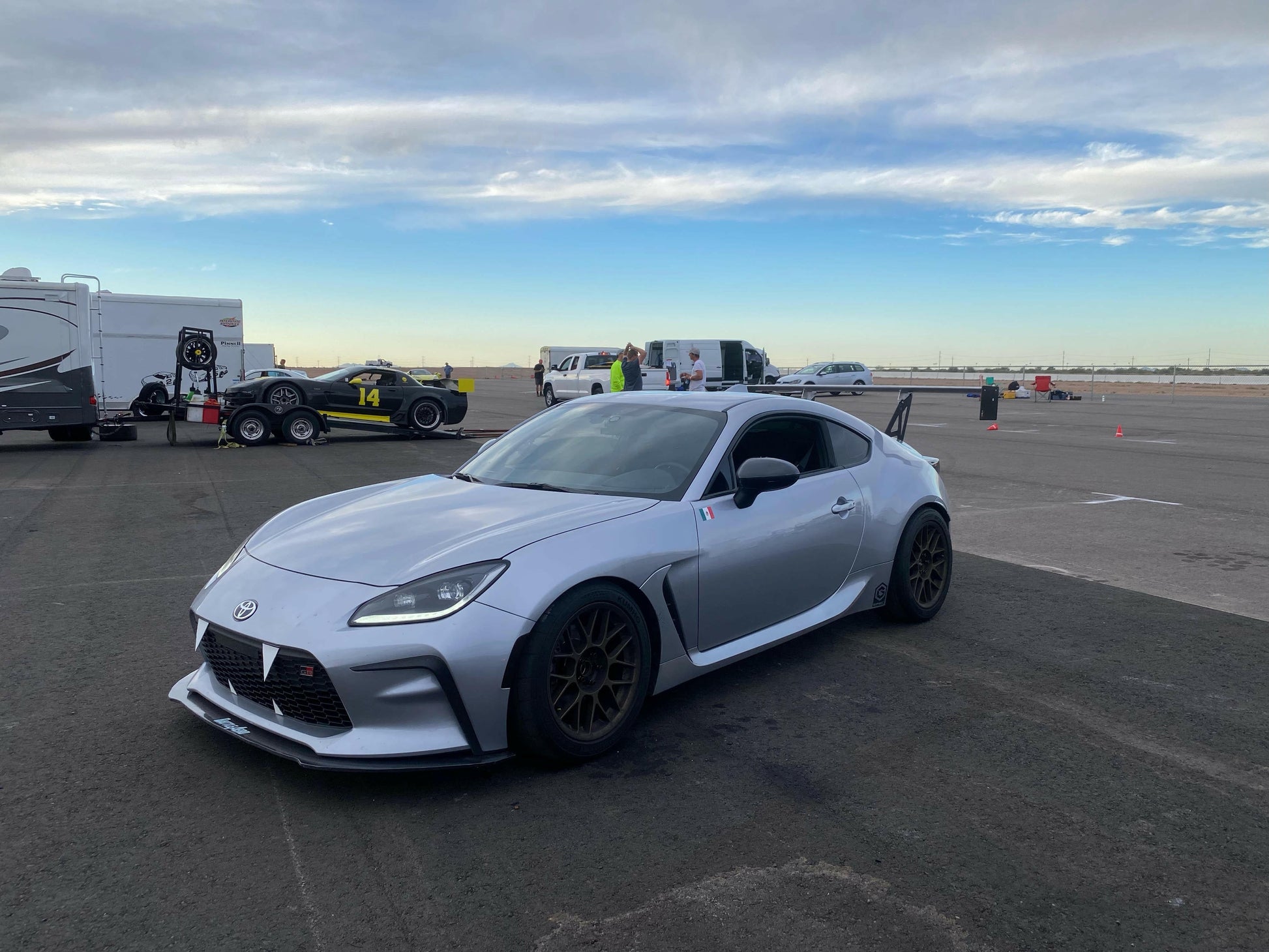 A silver sports car equipped with a 9livesracing GR86 / BRZ Wang Kit '22+, boasting extruded aluminum wings, is parked on a racetrack next to a black race car sporting the number 14. Several people and vehicles are in the background under a cloudy sky, creating an atmosphere of what seems to be a motorsport event or track day.