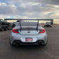 A silver Toyota sports car, equipped with the 9livesracing GR86/BRZ Wang Kit '22+ featuring large extruded aluminum wings that enhance its lift-to-drag ratio, is parked on an asphalt surface. Its license plate reads "POORSH" and displays an Arizona state emblem. In the background, another sports car is on a trailer alongside several other vehicles and a building under a cloudy sky.