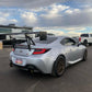 A silver Toyota sports car, equipped with a GR86 / BRZ Wang Kit '22+ from 9livesracing, featuring performance airfoils on its large rear spoiler, is parked in a lot. The car has a custom license plate reading "PROBRO." In the background under a partly cloudy sky, an RV and a white SUV are visible, along with two people standing near the SUV.